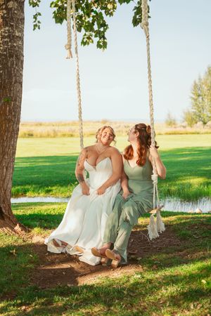 Two people in formal wear sit together on a rustic tree swing in a scenic outdoor setting during golden hour.