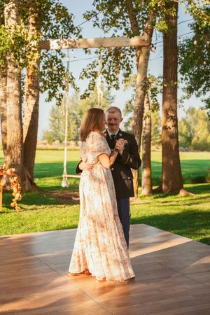 Couple dancing on wooden platform beneath string lights and birch trees during sunset.