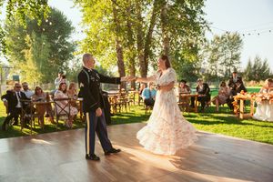 A couple shares their first dance outdoors on a wooden dance floor at a sunset wedding reception with guests looking on.
