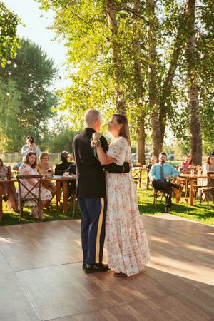 Couple shares their first dance on outdoor wooden dance floor at sunset wedding reception with guests watching from tables.