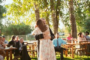 Wedding guests enjoying an outdoor reception under trees with wooden picnic tables in a sunlit garden setting.