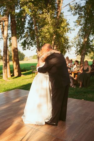 A couple shares a romantic dance moment during sunset on an outdoor wooden dance floor with trees in the background.