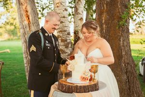 A couple cutting their rustic wedding cake displayed on a wood slice stand beneath birch trees.