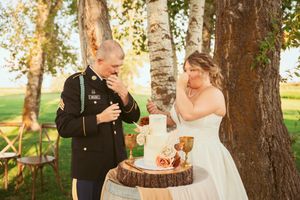 Military couple performs wedding cake cutting ceremony outdoors near birch trees.