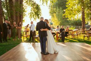 A couple shares their first dance on an outdoor dance floor surrounded by guests and string lights.