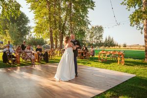 A couple dances on an outdoor wooden floor with guests seated at tables under tall trees.