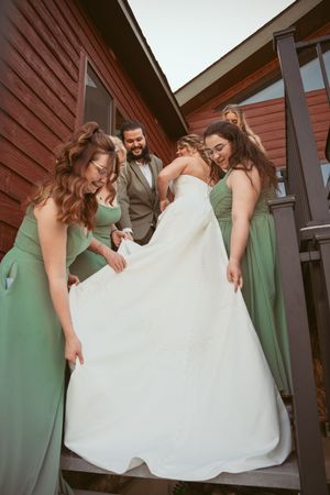 Wedding party in sage green dresses helps adjust a flowing white wedding gown on outdoor steps.