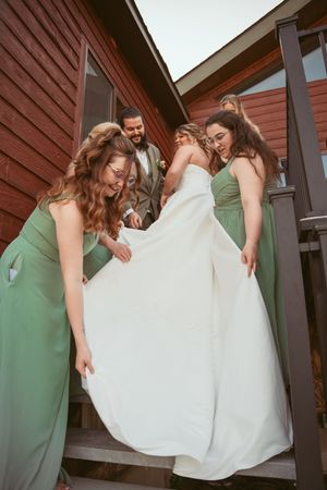Bridesmaids in sage green dresses help adjust a white wedding gown on outdoor wooden stairs.