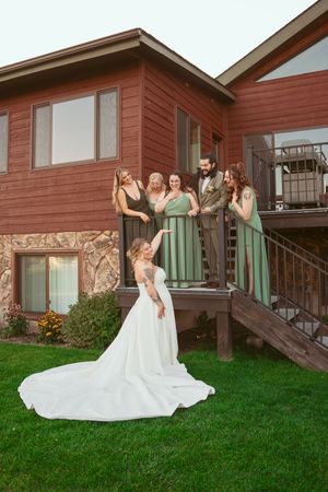 A wedding party gathered on rustic house steps with bridesmaids in sage green dresses and a flowing white bridal gown.