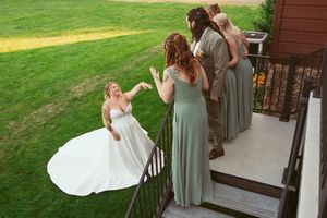 Bridesmaids in sage green dresses gather on a porch with a bride in a flowing white gown on outdoor stairs.
