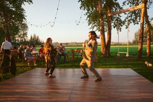 A lively outdoor dance scene on a wooden deck with string lights and trees at sunset in a rural setting.