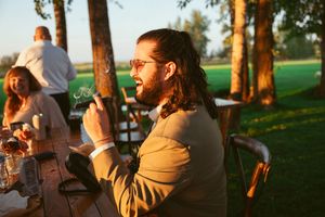 Person in brown jacket sits outdoors at a table during golden hour in a park setting with trees in background.