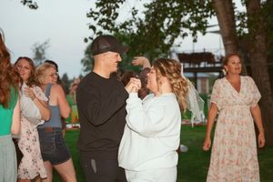 A group of people dance outdoors on grass at dusk with trees and a gazebo visible in the background.