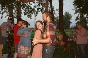 People dancing and embracing at an outdoor party with trees and picnic tables in the background at dusk.