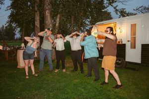 A group of people dance outdoors at dusk in front of a white RV trailer with warm lighting in the background.