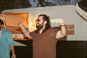 Person wearing brown sweater holding beverage at outdoor party with festive lighting in background.