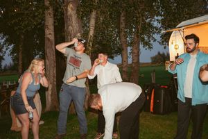 A group of friends share a casual moment outdoors at dusk, laughing and drinking while gathered near trees.