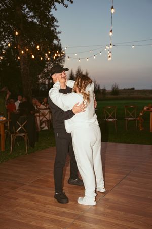A couple shares their first dance on a wooden dance floor under romantic string lights at dusk.