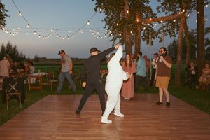 People dancing on an outdoor wooden deck under string lights at dusk with trees and farmland in the background.