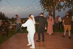 Wedding guests dancing and enjoying a reception on an outdoor wooden deck with string lights overhead at dusk.