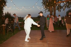 Party guests dance on a wooden deck under twinkling lights with trees in the background at sunset.