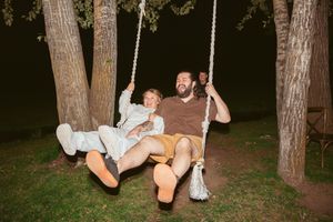 A rustic outdoor swing decorated with string lights hangs between two pine trees at dusk.