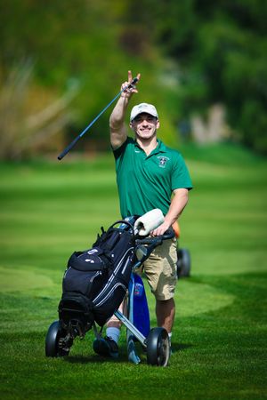 A golfer in a green polo gives a thumbs up while walking with a golf bag cart on a lush fairway.