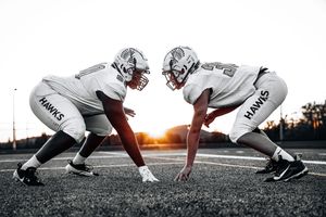 Two football players in white uniforms facing each other in three-point stance at sunset.