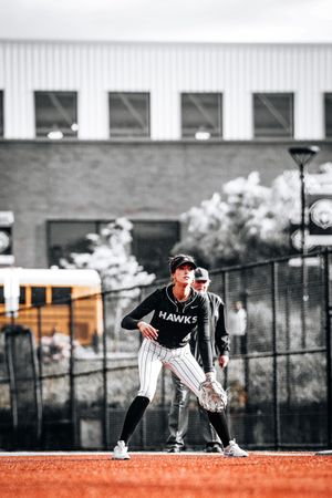 Baseball player in Hawks jersey crouched in fielding position on orange-accented field against monochrome backdrop.