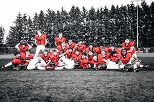 Black and white photo with red selective coloring shows youth football team posing in uniform on grass field with pine trees.