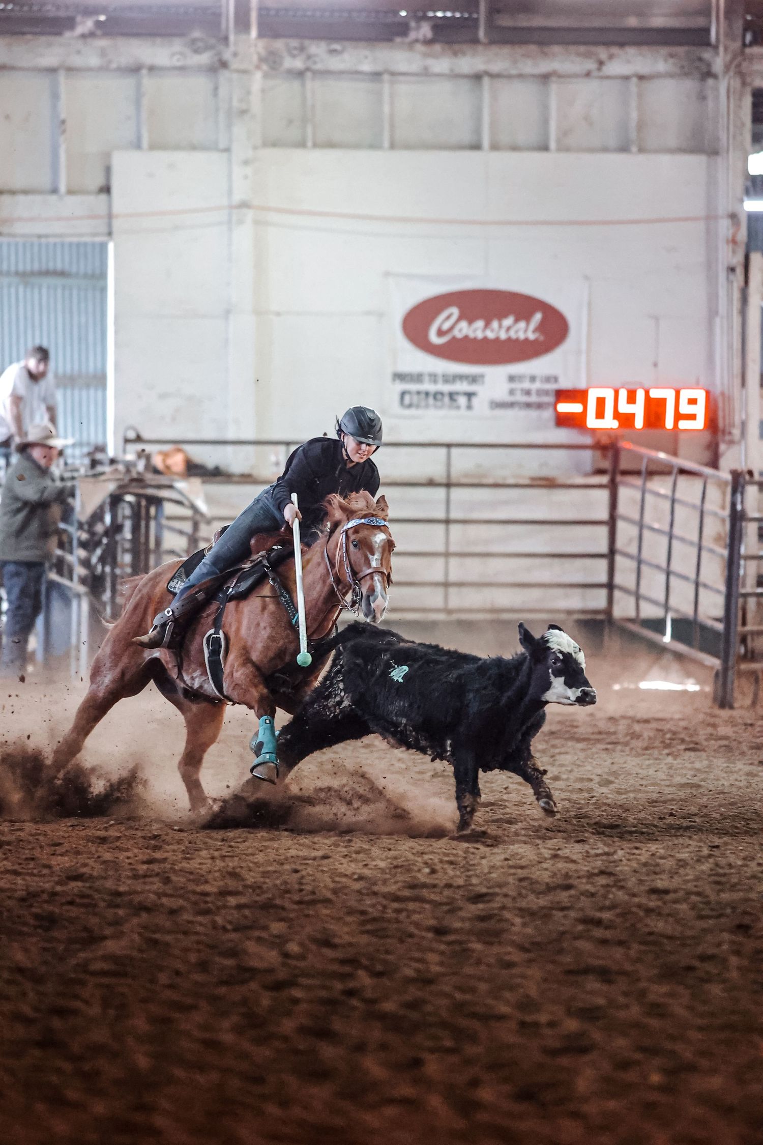 A cowboy on horseback ropes a calf during a rodeo competition in an indoor dusty arena with a digital timer.