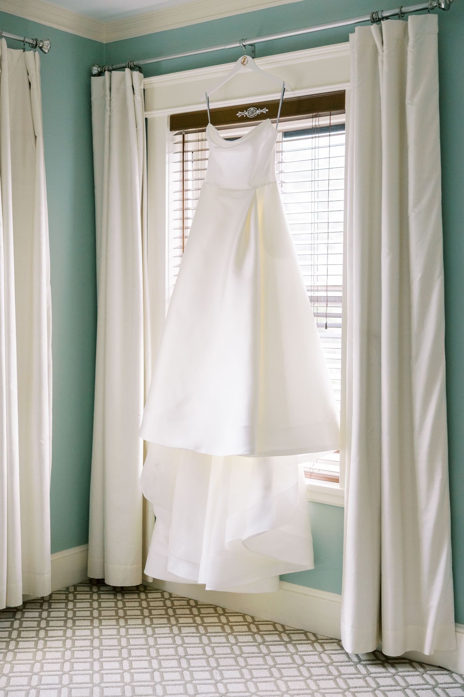 An elegant white wedding dress hangs by a window with cream curtains in a mint green room with patterned tile floors.