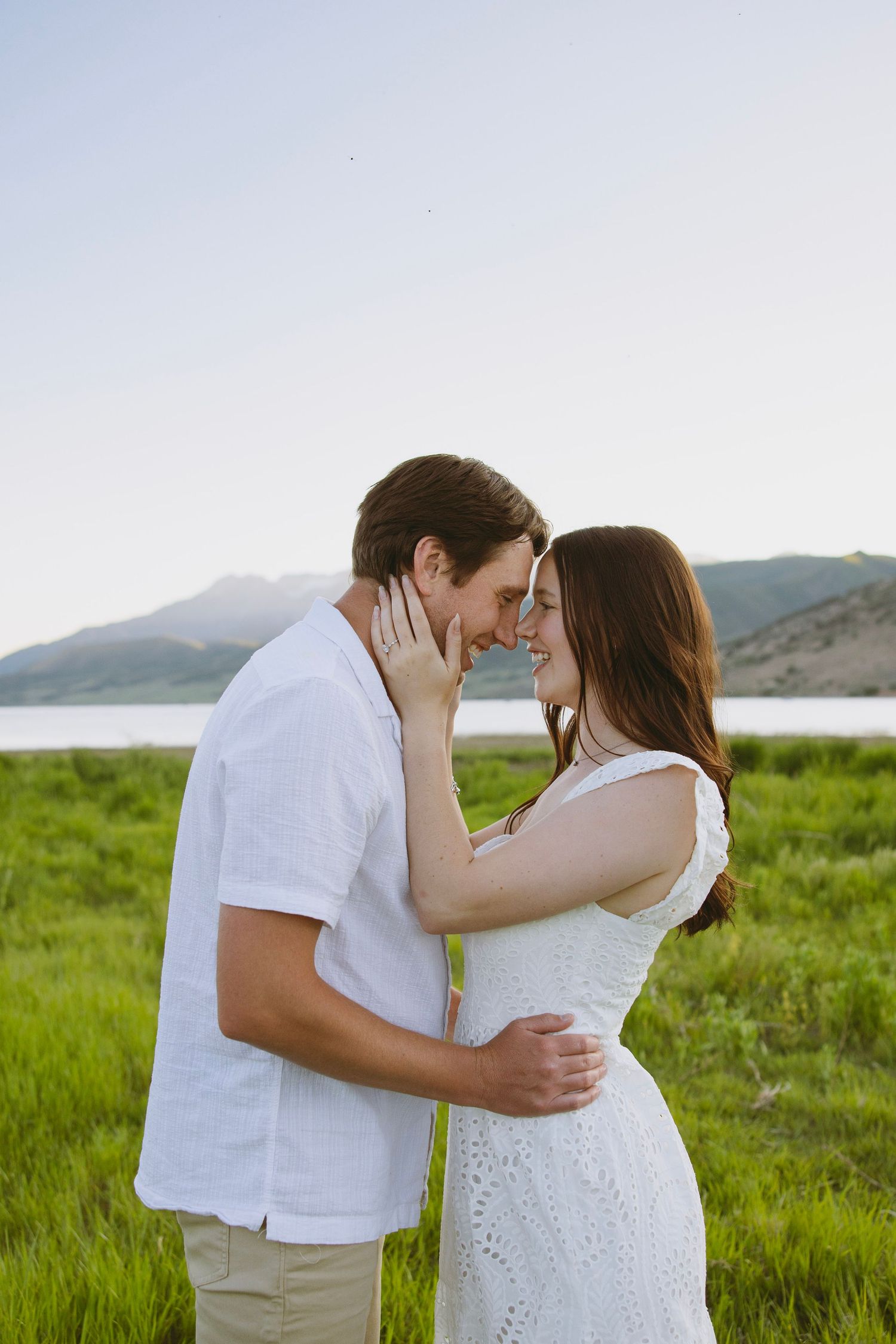 Bride in flowing white dress holds bouquet while standing on wooden deck surrounded by evergreen mountains at sunset.