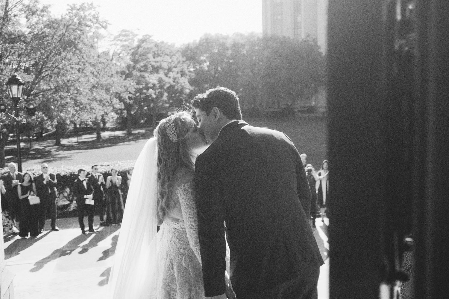 A black and white wedding photo shows a couple sharing a romantic moment on church steps as guests gather below.