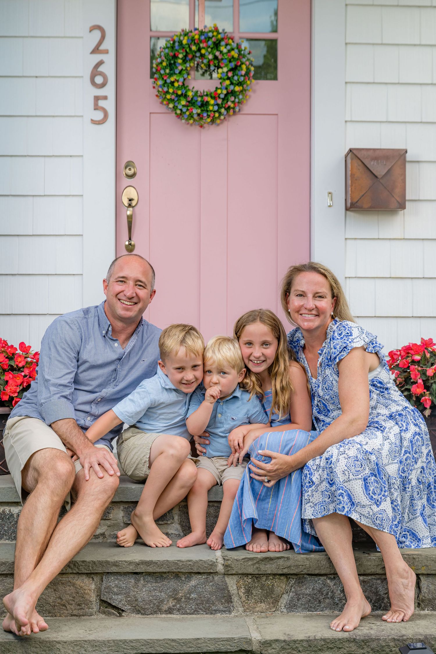 A family sits barefoot on stone steps in front of a pink door with a wreath and red flowers blooming nearby.