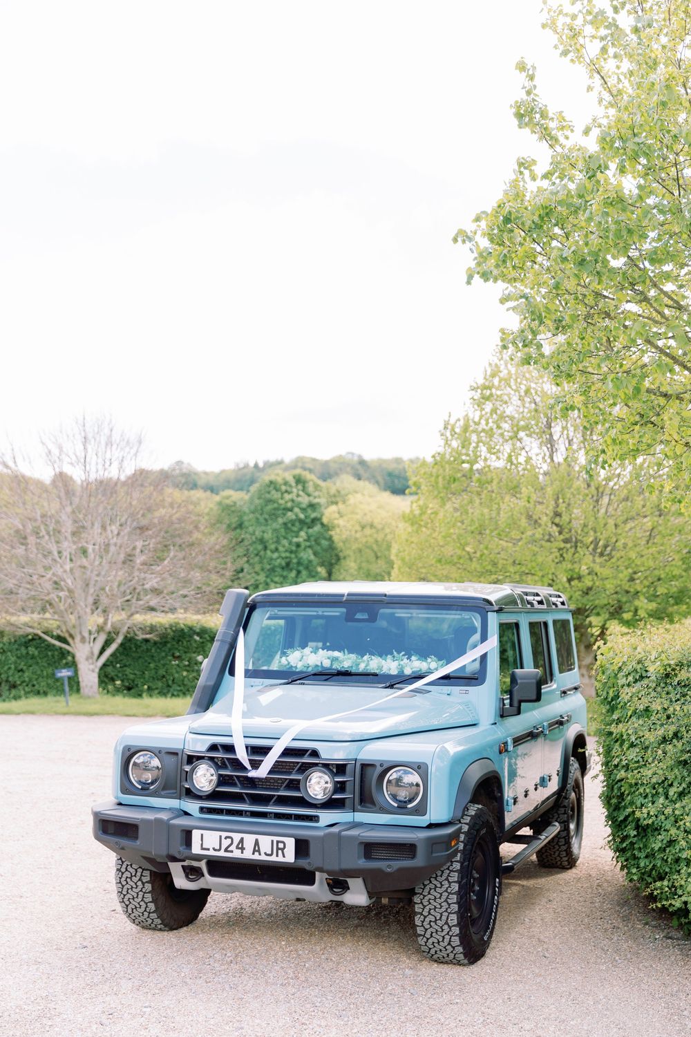Light blue Land Rover Defender decorated with wedding ribbons parked in countryside setting.
