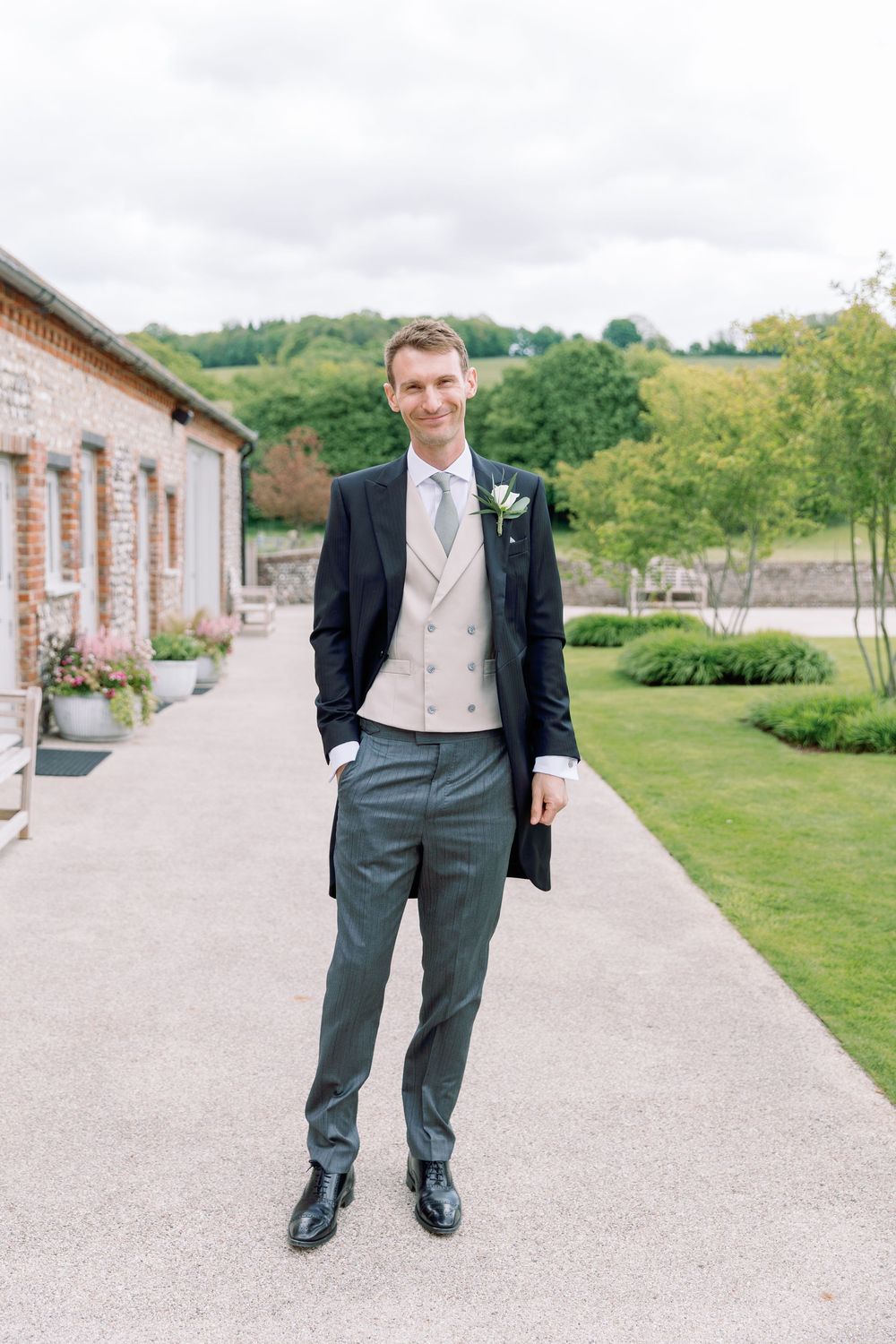Groom in classic morning suit with tails and vest posing on pathway outside wedding venue.