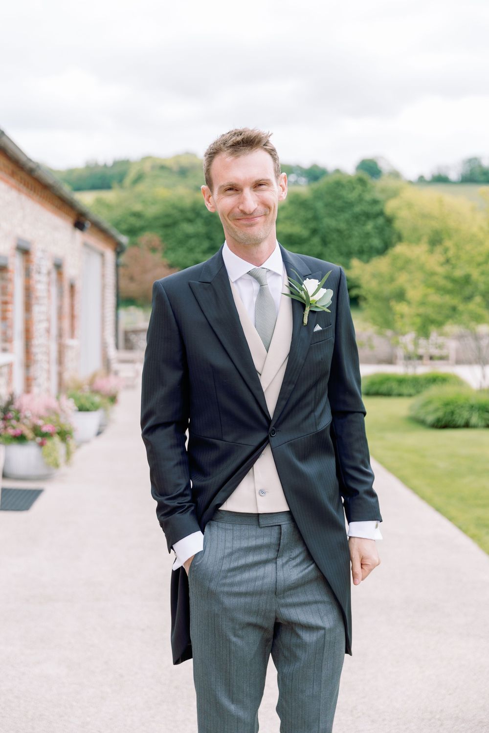 Groom in traditional morning suit with boutonniere smiling on venue grounds.