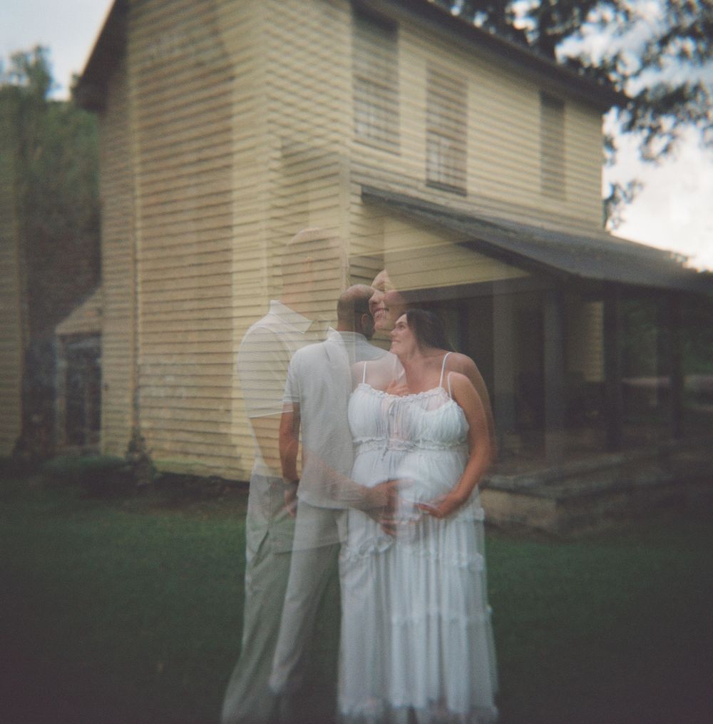 A couple in white attire stands together in front of a rustic two-story farmhouse on a grassy lawn at dusk.