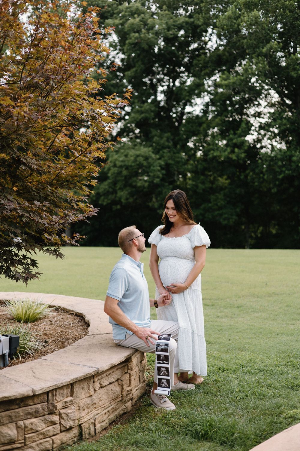 Couple shares tender moments on stone wall during outdoor maternity photo session in park setting with autumn trees.