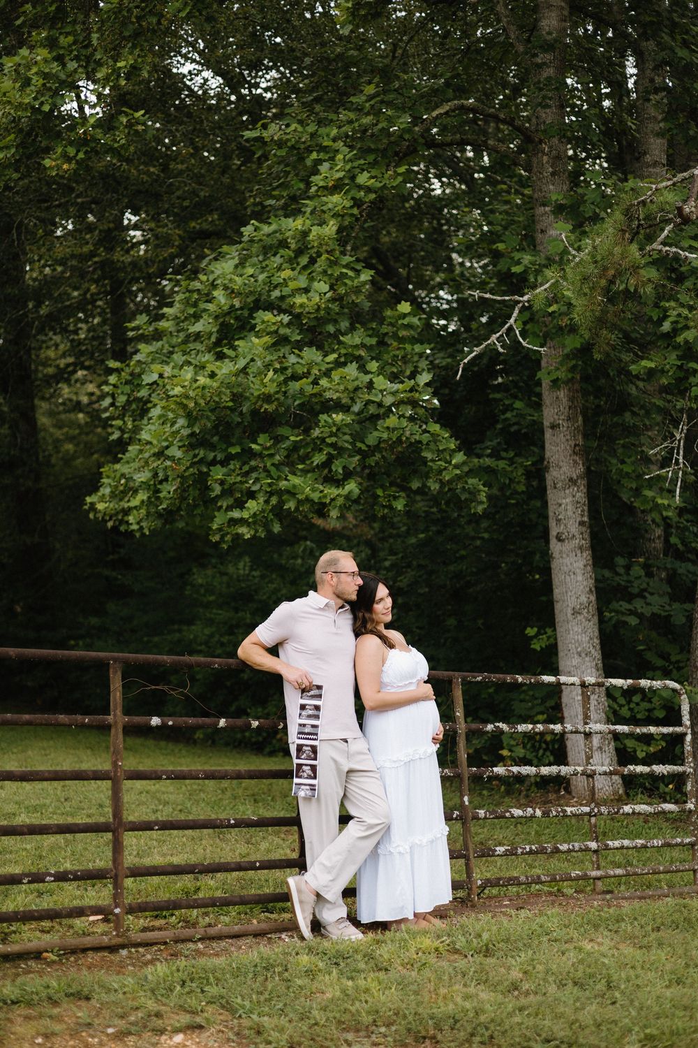 A couple in white outfits embraces while standing by a rustic fence in a wooded setting.