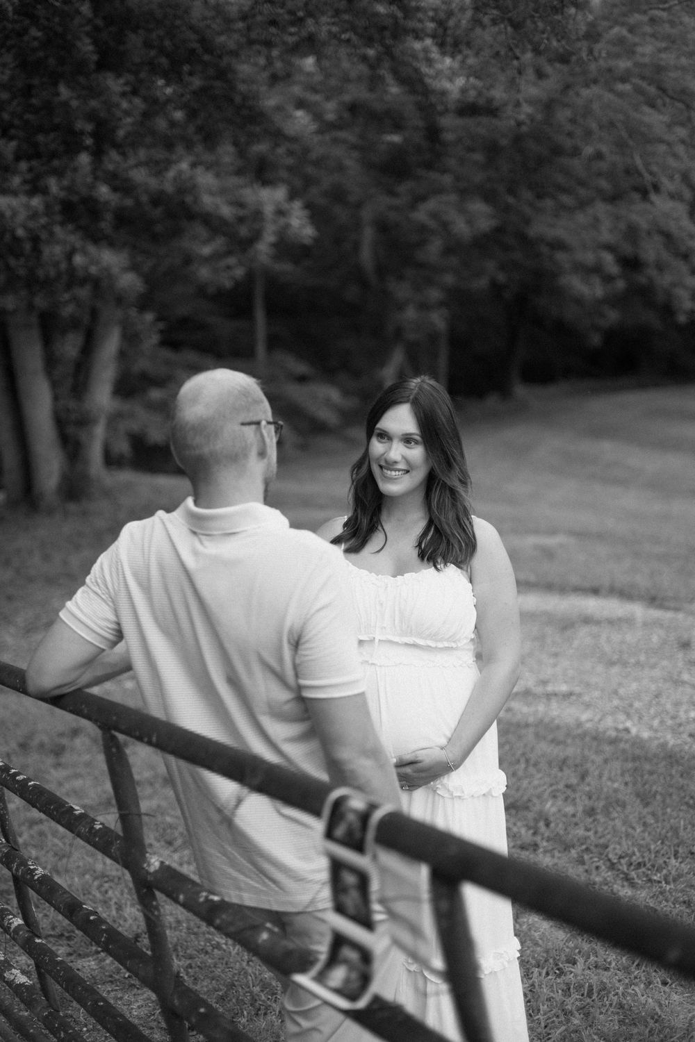 A couple sits together on a rustic wooden fence surrounded by trees in a black and white photograph.
