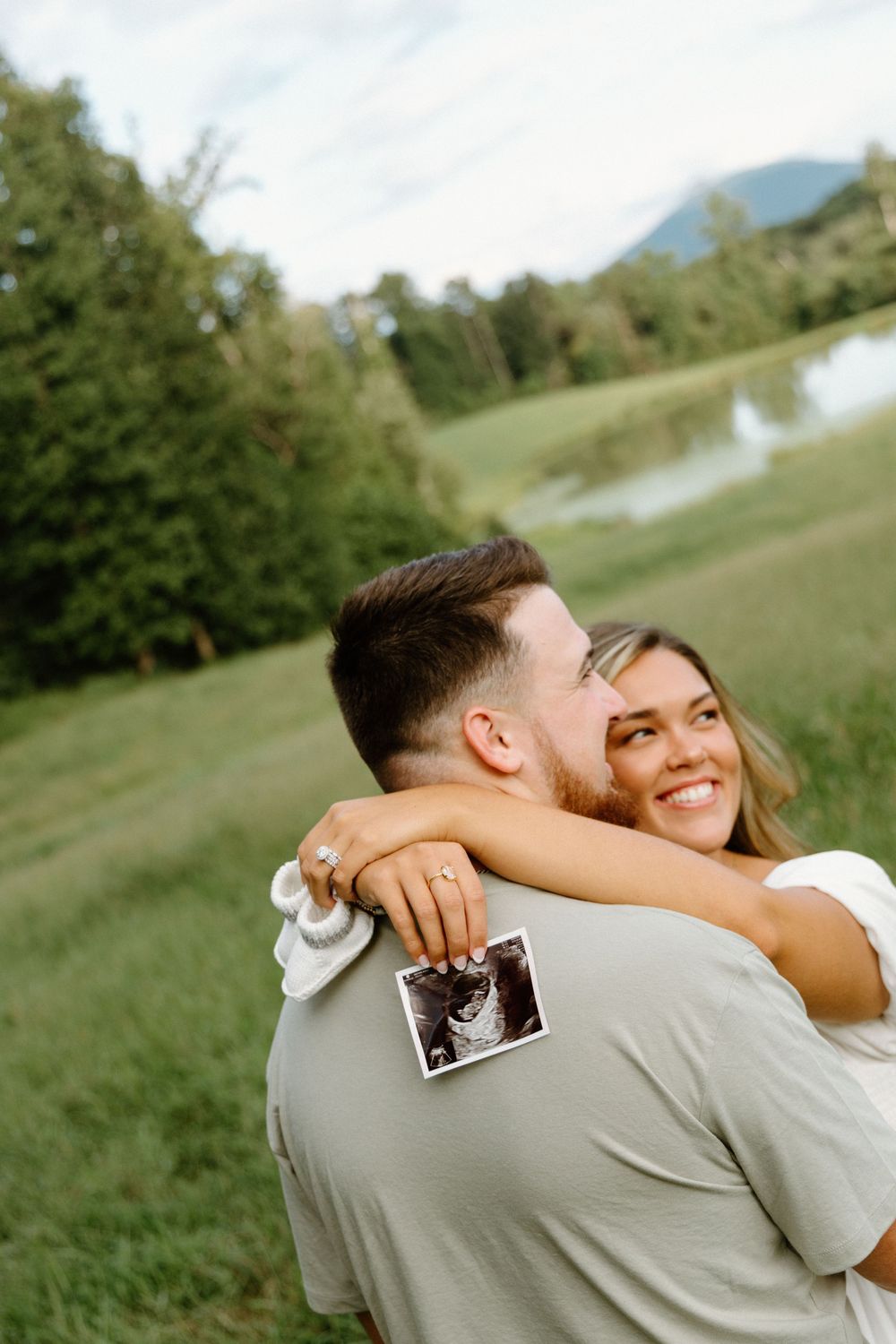A couple embraces outdoors while holding a sonogram photo during a pregnancy announcement moment.