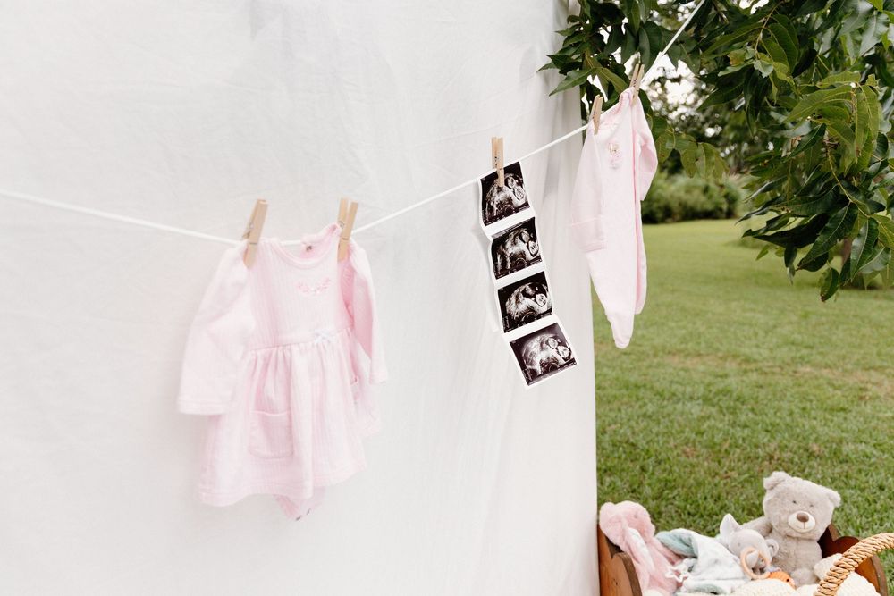 A collection of pink onesies and baby clothes hung on a clothesline outdoors with ultrasound photos in between them.