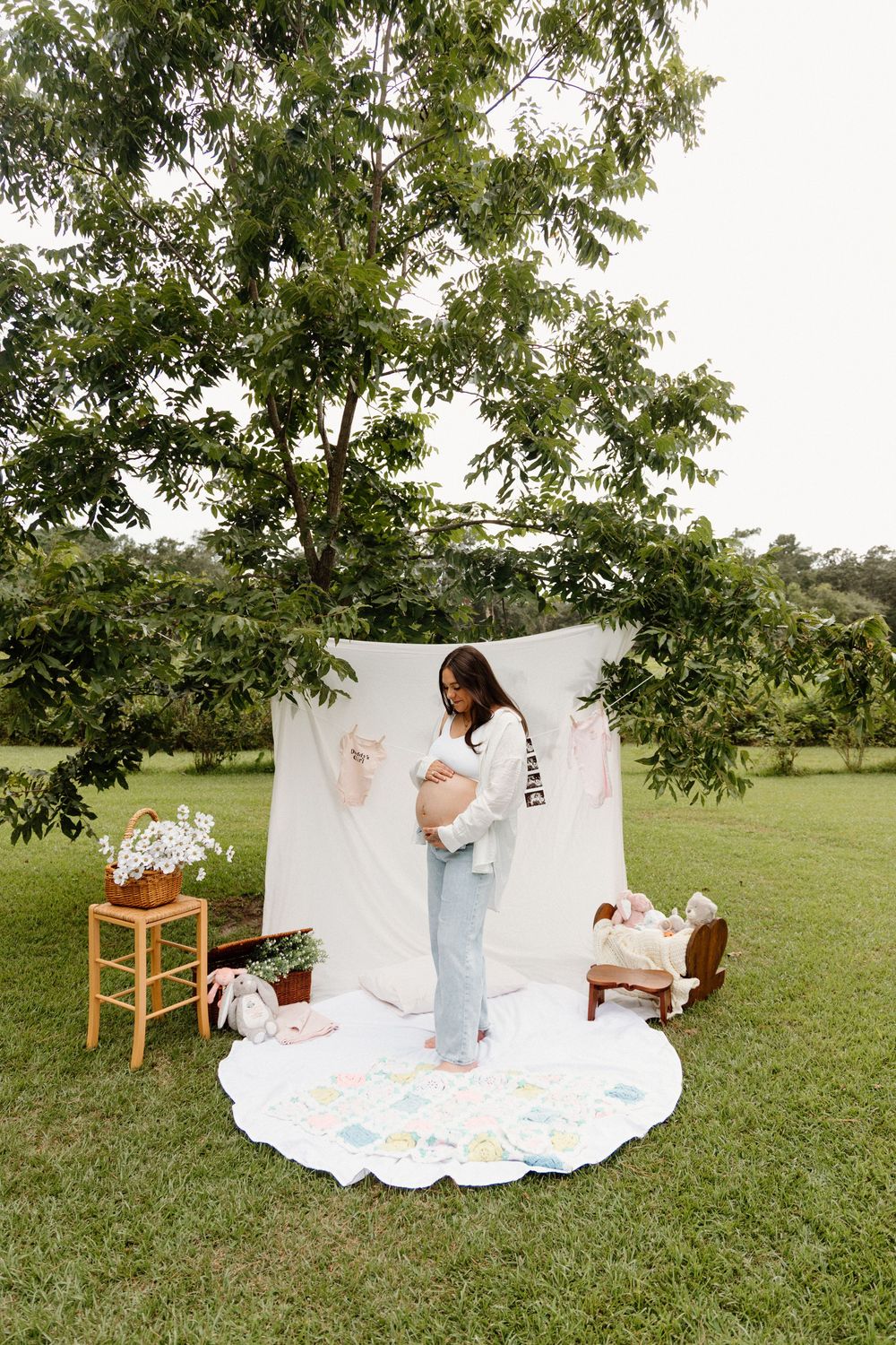White blanket spread out on green grass under tree with wooden chair setup for outdoor photography session.