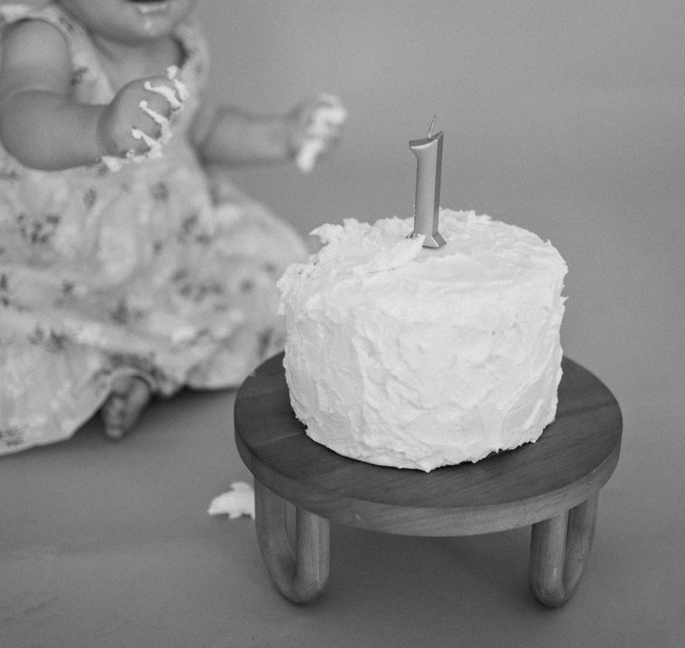 A white frosted smash cake with number one candle sits on a wooden cake stand in black and white photography.