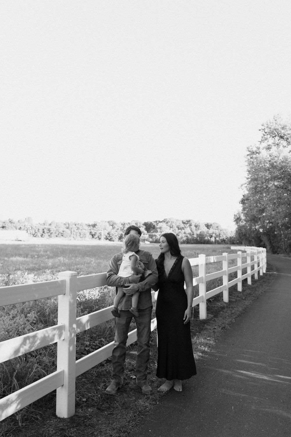 Black and white photo of a couple with a baby standing by a white fence overlooking a scenic countryside view.