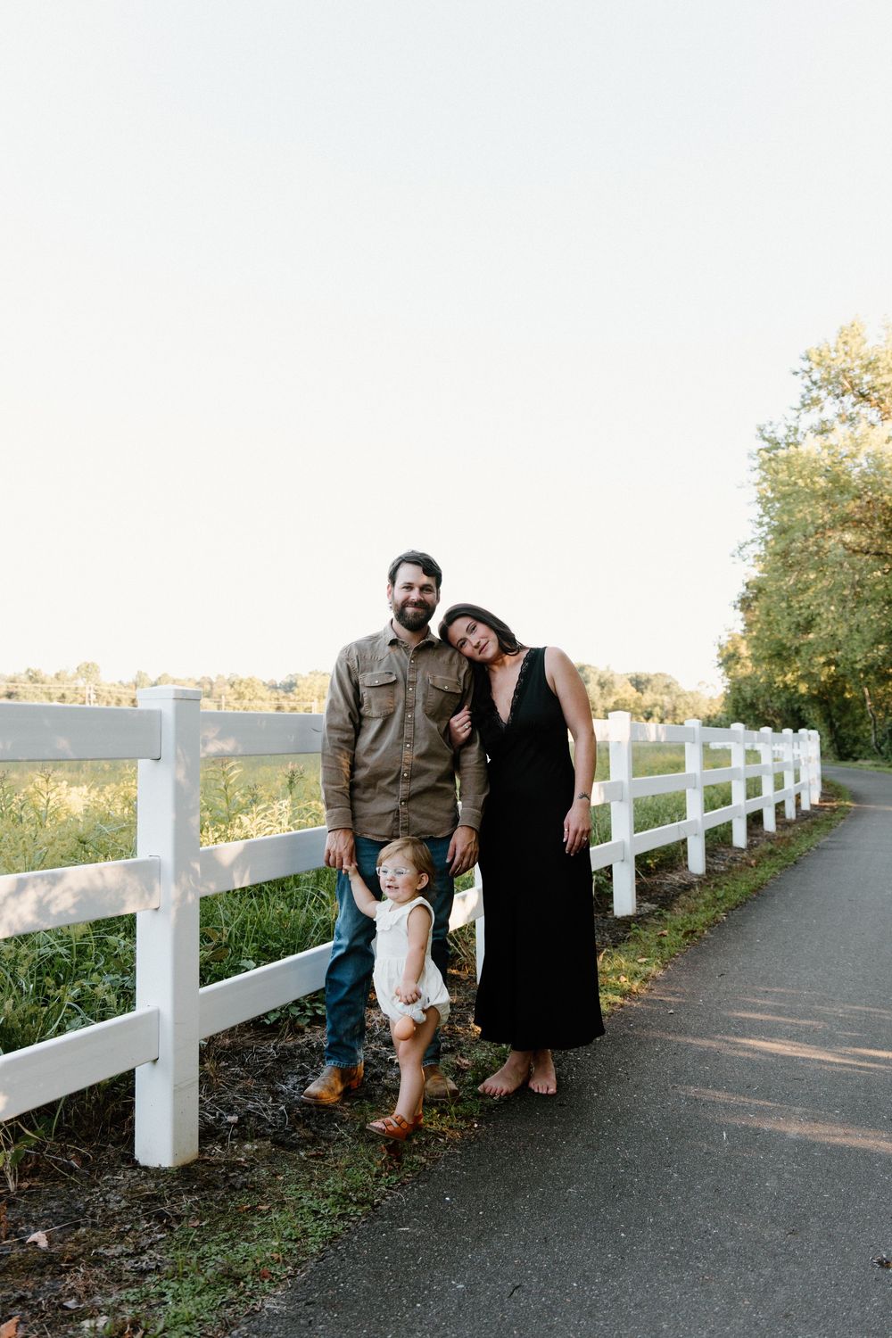 A couple sits together on a white fence-lined path during golden hour on an autumn day.