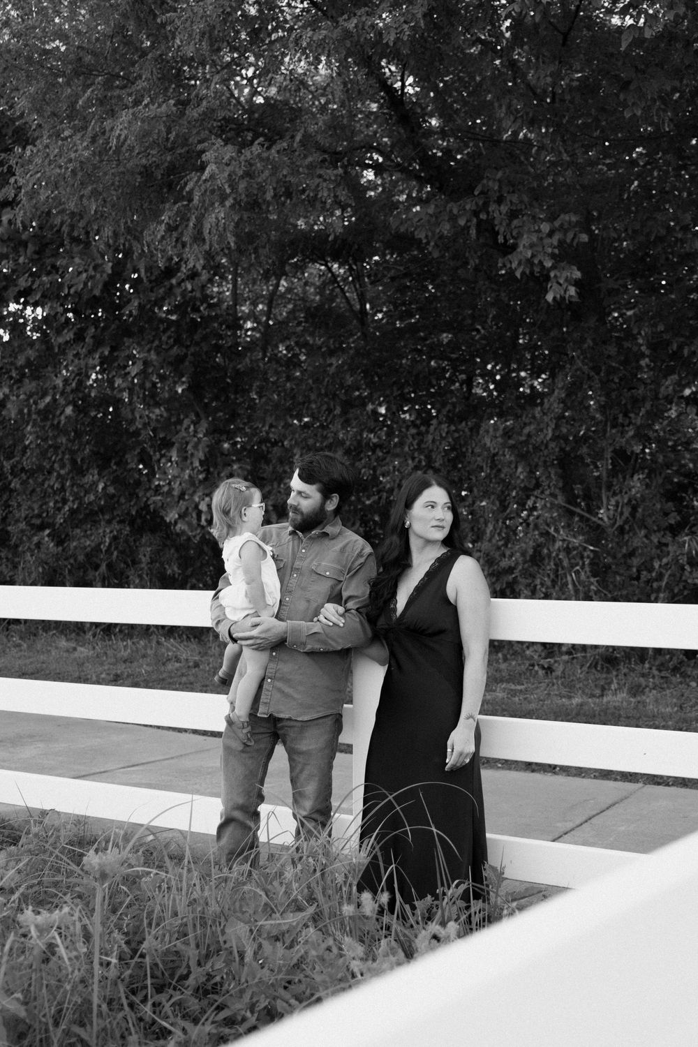 A couple shares an intimate moment sitting together on a white fence while surrounded by foliage in black and white.