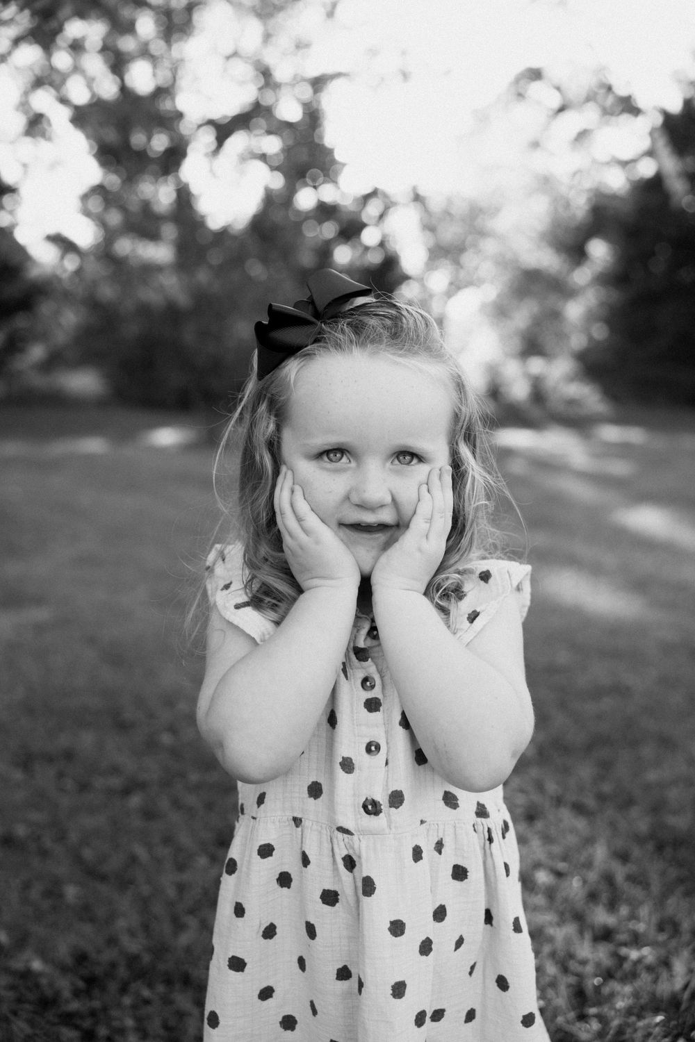 A child in a polka dot dress plays peek-a-boo outdoors in black and white photography.